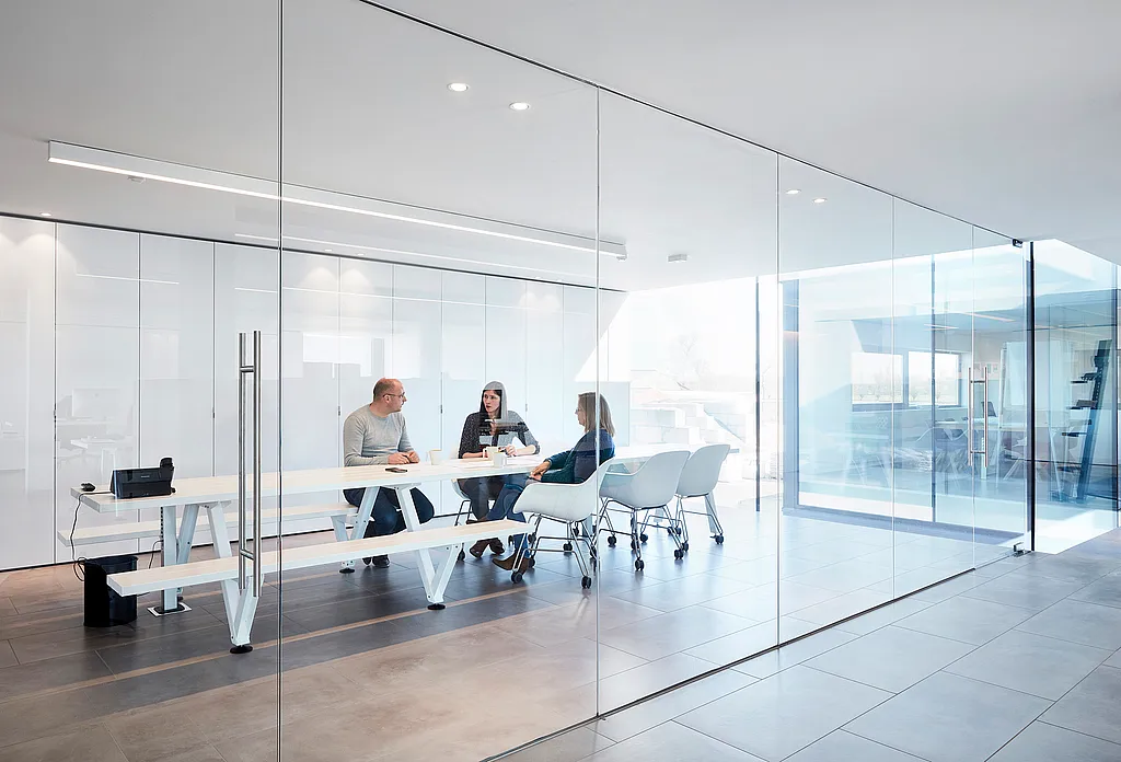 Minimale vergaderruimte met glazen wanden met een lange witte Marina Combo-tafel en bijpassende bank, waar drie collega's samen op witte Captain's Chairs zitten in een licht modern kantoor.