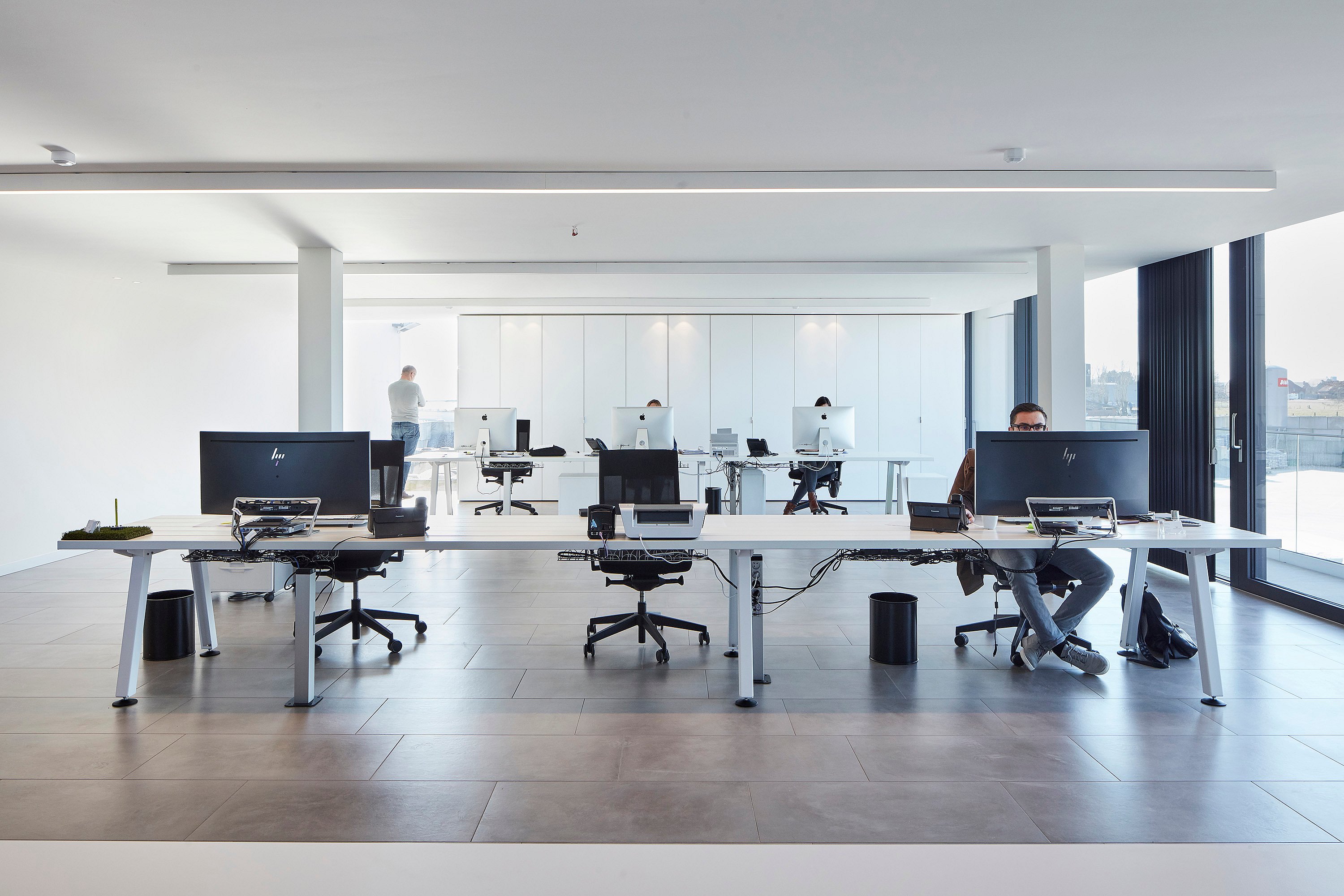 Helles, offenes Büro mit langen weißen Marina-Schreibtischen, großen Computerbildschirmen und raumhohen Fenstern mit Blick auf die Terrasse.