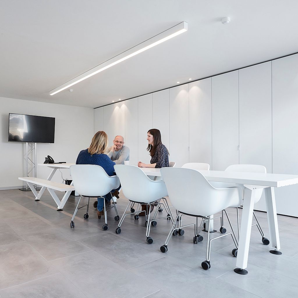 Drei Kollegen unterhalten sich an einem langen weißen Marina-Kombitisch in einem hellen Konferenzraum, der mit hohen weißen Schränken und weißen Kapitänsstühlen auf Rollen gesäumt ist.