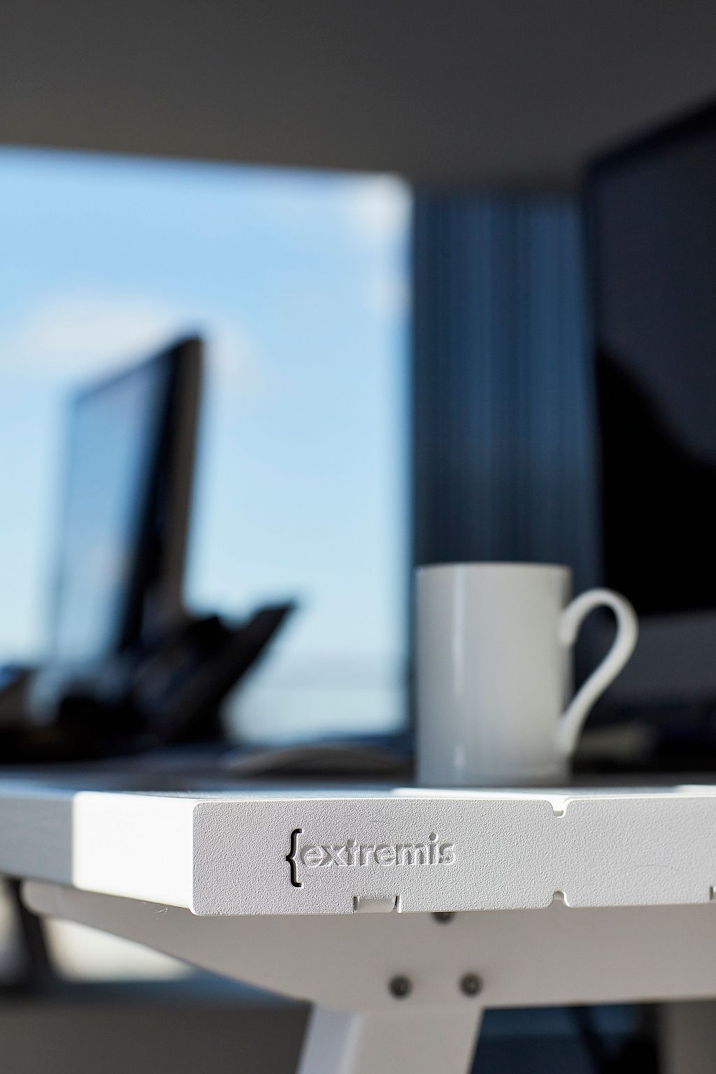 Nahaufnahme der Ecke eines weißen Marina-Tisches mit dem geprägten Extremis-Logo, einer weißen Tasse und unscharfen Bürobildschirmen im Hintergrund.