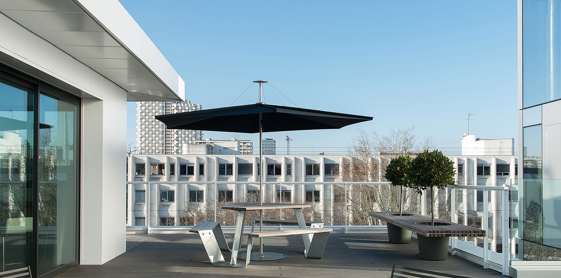 Weißer Anker-Picknicktisch unter einem dunklen Inumbrina-Sonnenschirm auf einer geräumigen Dachterrasse mit einer modernen weißen Gebäudefassade und der Skyline von Rennes im Hintergrund.
