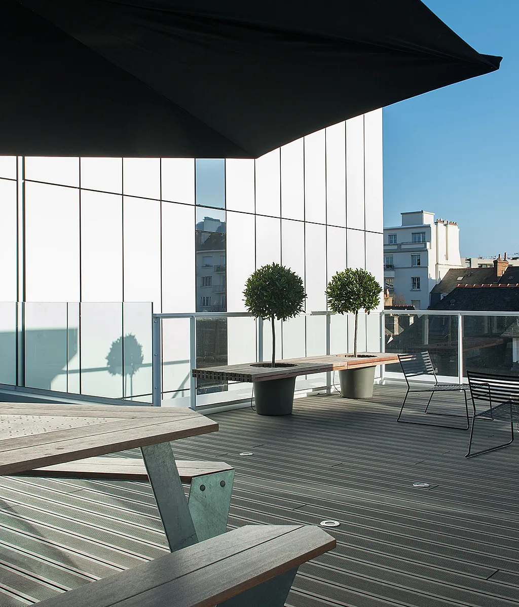 Moderne Büroterrasse auf dem Dach mit Anker-Picknicktischen und großen dunklen Inumbrina-Sonnenschirmen, gesäumt von einem Glasgeländer und vor einem weißen, modernen Gebäude mit Blick auf die Stadt dahinter.