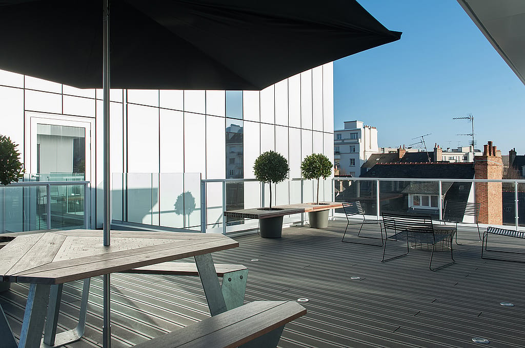 Moderne Büroterrasse auf dem Dach mit Anker-Picknicktischen und großen dunklen Inumbrina-Sonnenschirmen, gesäumt von einem Glasgeländer und vor einem weißen, modernen Gebäude mit Blick auf die Stadt dahinter.