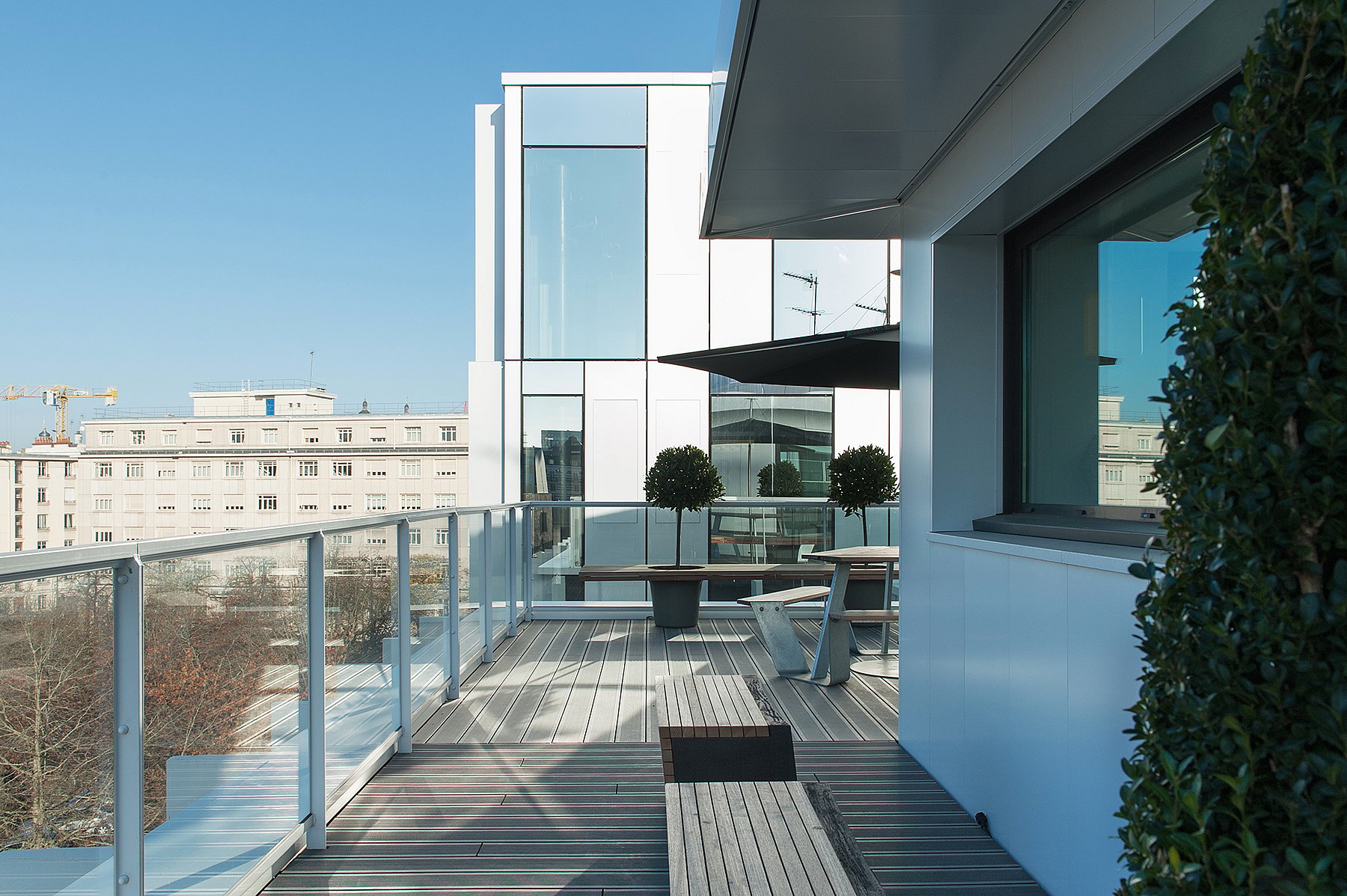 Glasgefütterte Büroterrasse mit Anker-Sitzen, runden Formschnittbäumen in Romeo & Juliet-Pflanzgefäßen und einem dunklen Sonnenschirm, der Schatten über das Lattendeck neben dem Gebäude wirft.