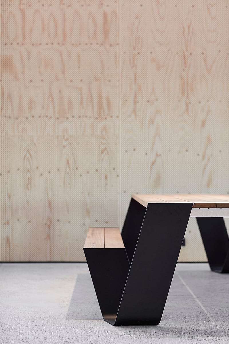 Multifunctional communal tables set within the open-plan Hangar K coworking hub, blending warm wood surfaces with black frames and raw industrial materials.