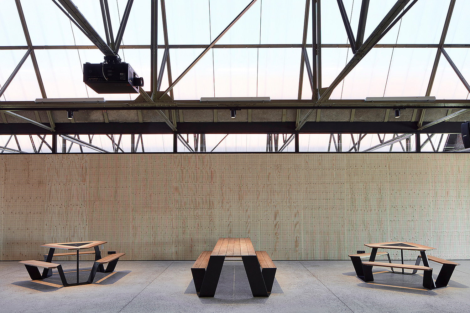 Industrial coworking space inside Hangar K with communal picnic tables in the foreground, glass-walled meeting rooms, and an open steel roof structure above.
