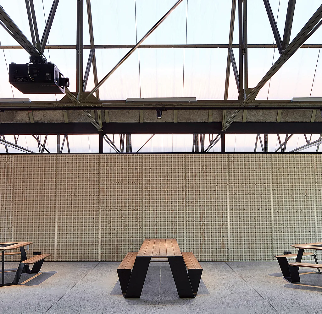 Industriële coworking-ruimte in Hangar K met gemeenschappelijke picknicktafels op de voorgrond, vergaderruimten met glazen wanden en een open stalen dakconstructie erboven.