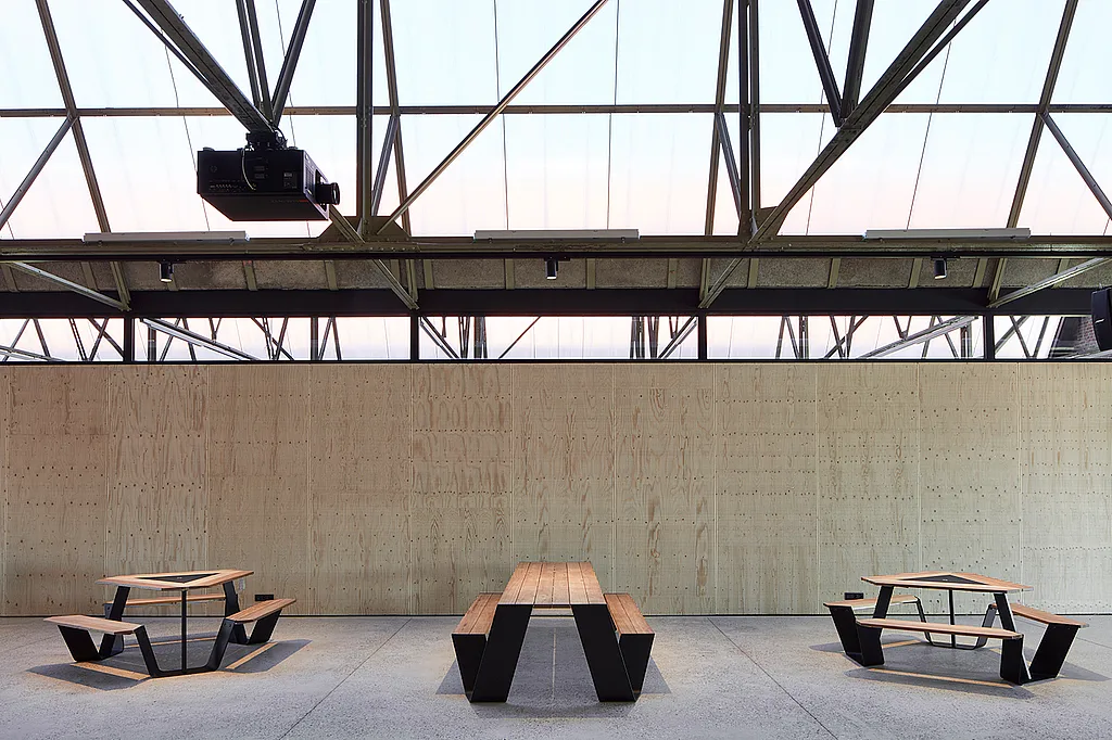 Industrial coworking space inside Hangar K with communal picnic tables in the foreground, glass-walled meeting rooms, and an open steel roof structure above.