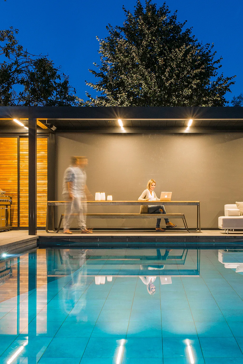 Wohnbereich im Freien neben einem Pool mit einem Hopper-Tisch unter einer modernen Aluminiumpergola mit Holzlatten, wodurch ein geschützter Raum zum Essen, Arbeiten und Entspannen entsteht.