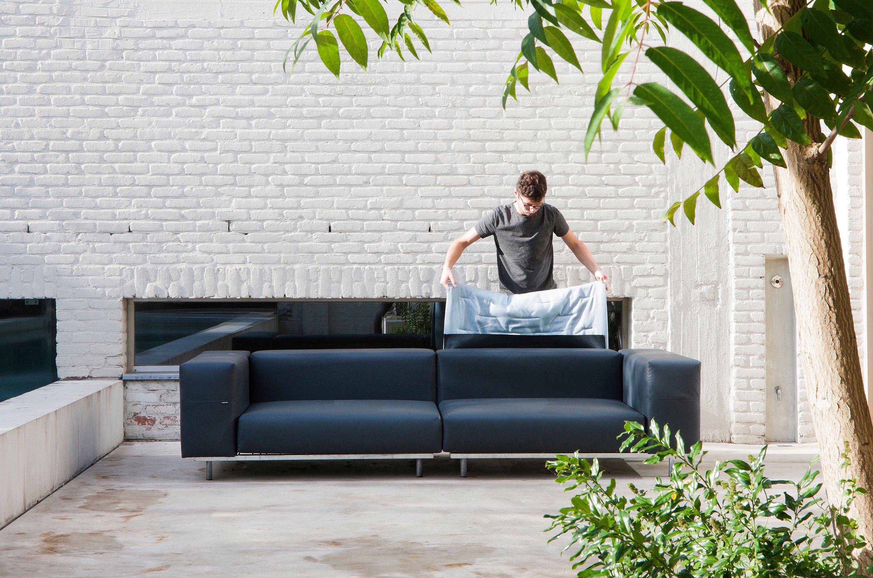 Man arranging a pale blue cushion on a dark gray Walrus sofa on a minimalist spa terrace with white brick walls and surrounding plants
