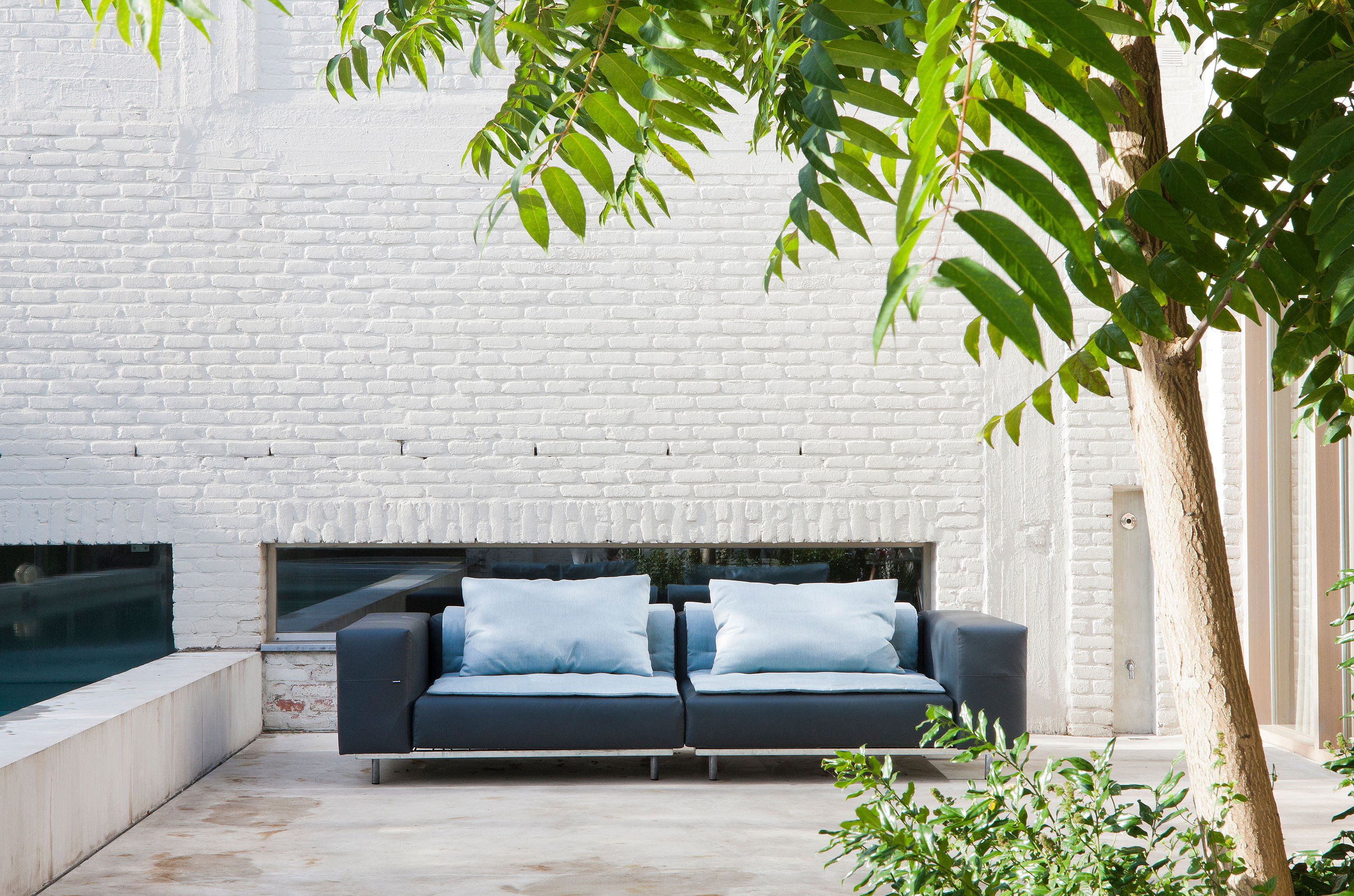 Dark gray Walrus sofa with two pale blue back cushions on a calm terrace beside a pool, set against a white-painted brick wall and framed by greenery.