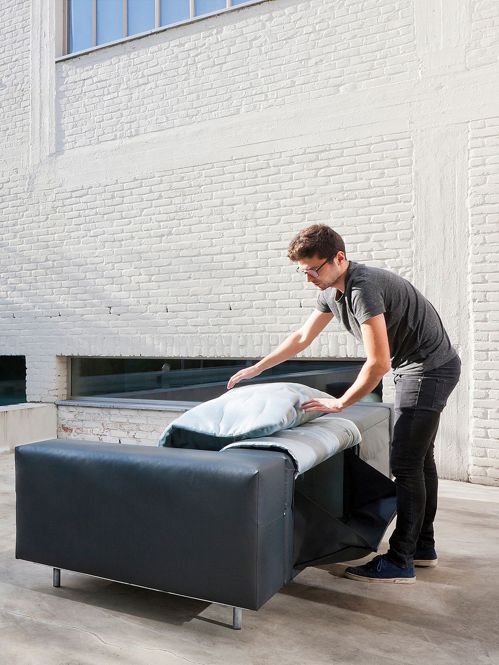 Man unfolding the light-colored cushion layer of a dark gray Walrus sofa, showing the flexible lounge setup on the spa terrace.