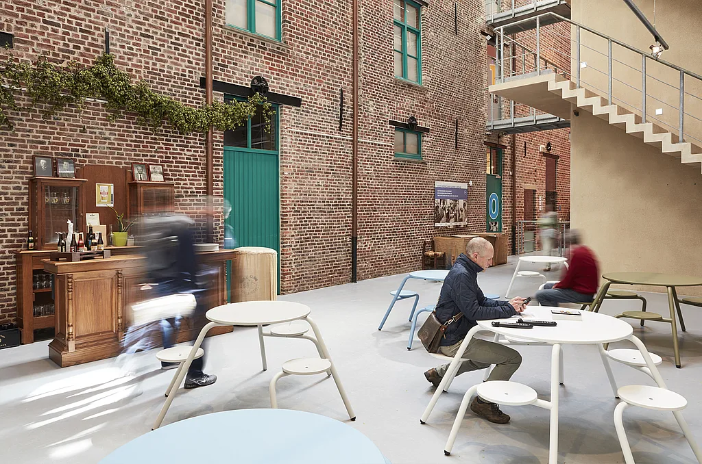 Historic brick museum courtyard with colorful round Virus tables, where visitors sit and move through the space beneath a metal staircase and teal-painted doors.