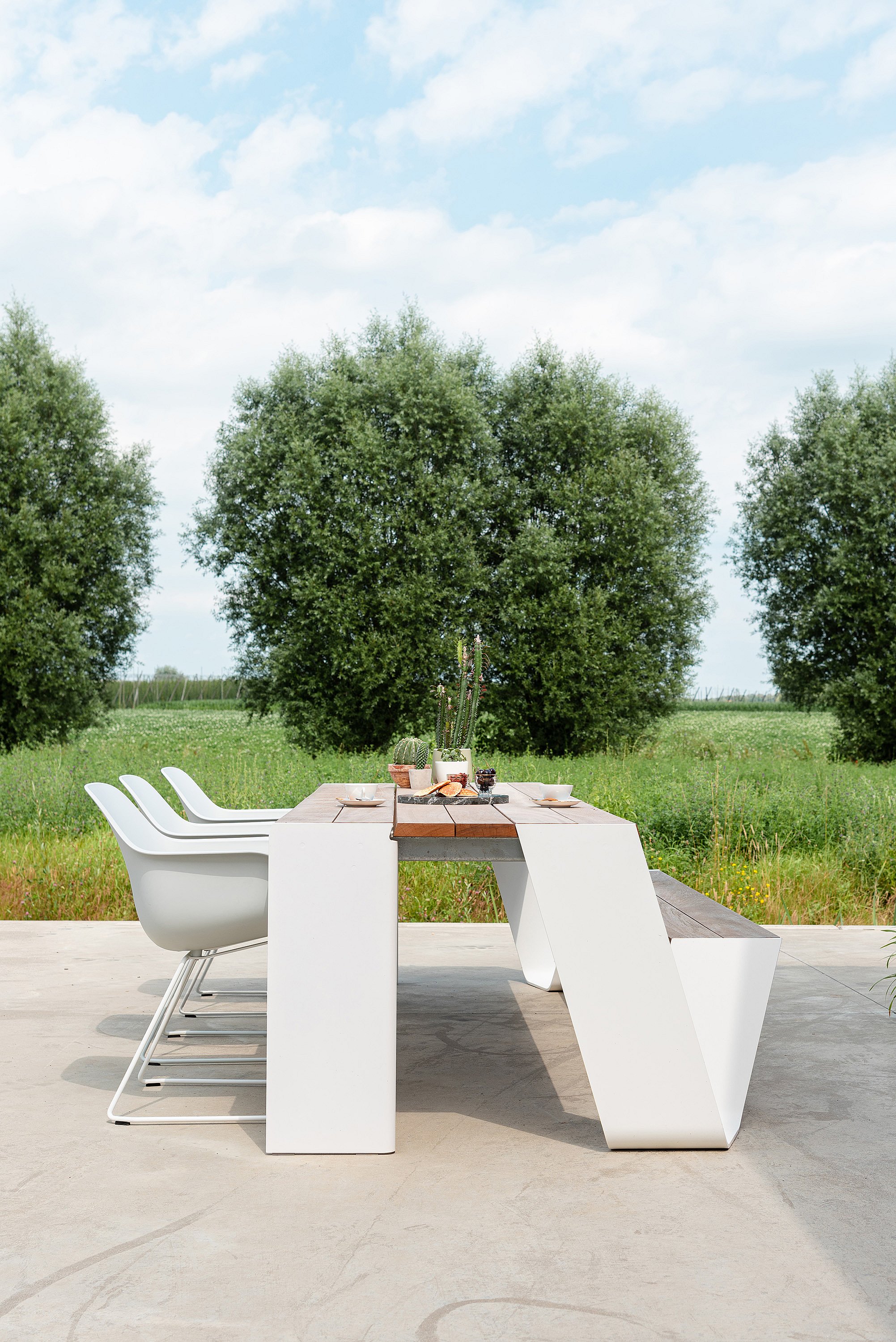 Vue de côté d’une table combo Hopper à l’extérieur, montrant trois chaises du capitaine blanches d’un côté et un banc en bois de l’autre, avec des arbres et une campagne ouverte derrière.