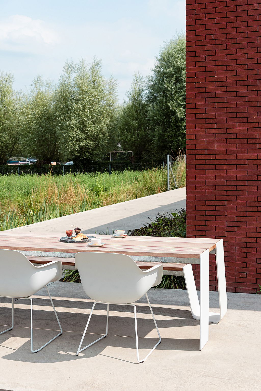 Longue table en bois pour l’extérieur avec des pieds blancs inclinés et des chaises du capitaine blanches, décorée avec des tasses à café et des pâtisseries sur une terrasse ensoleillée à côté d’un mur de briques rouges.