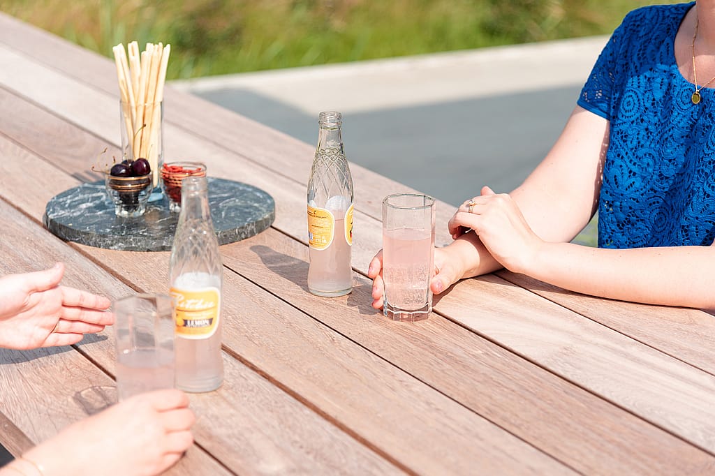 Gros plan d’une table en bois avec deux bouteilles de limonade, des verres transparents et un plateau de service rond contenant des cerises et des gressins sous un soleil éclatant.