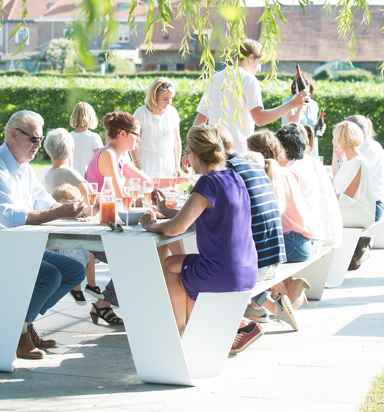Une grande famille s’est réunie autour de deux tables de pique-nique Hopper connectées sur une terrasse ensoleillée, partageant des boissons et de la nourriture sous les branches de saule suspendues.