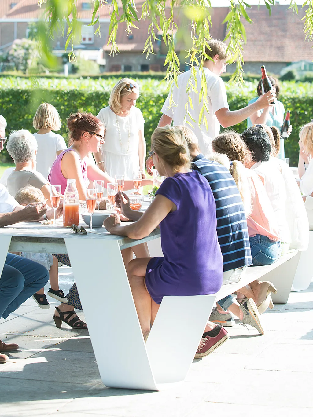Large family gathered around two connected Hopper picnic tables on a sunny patio, sharing drinks and food beneath hanging willow branches.