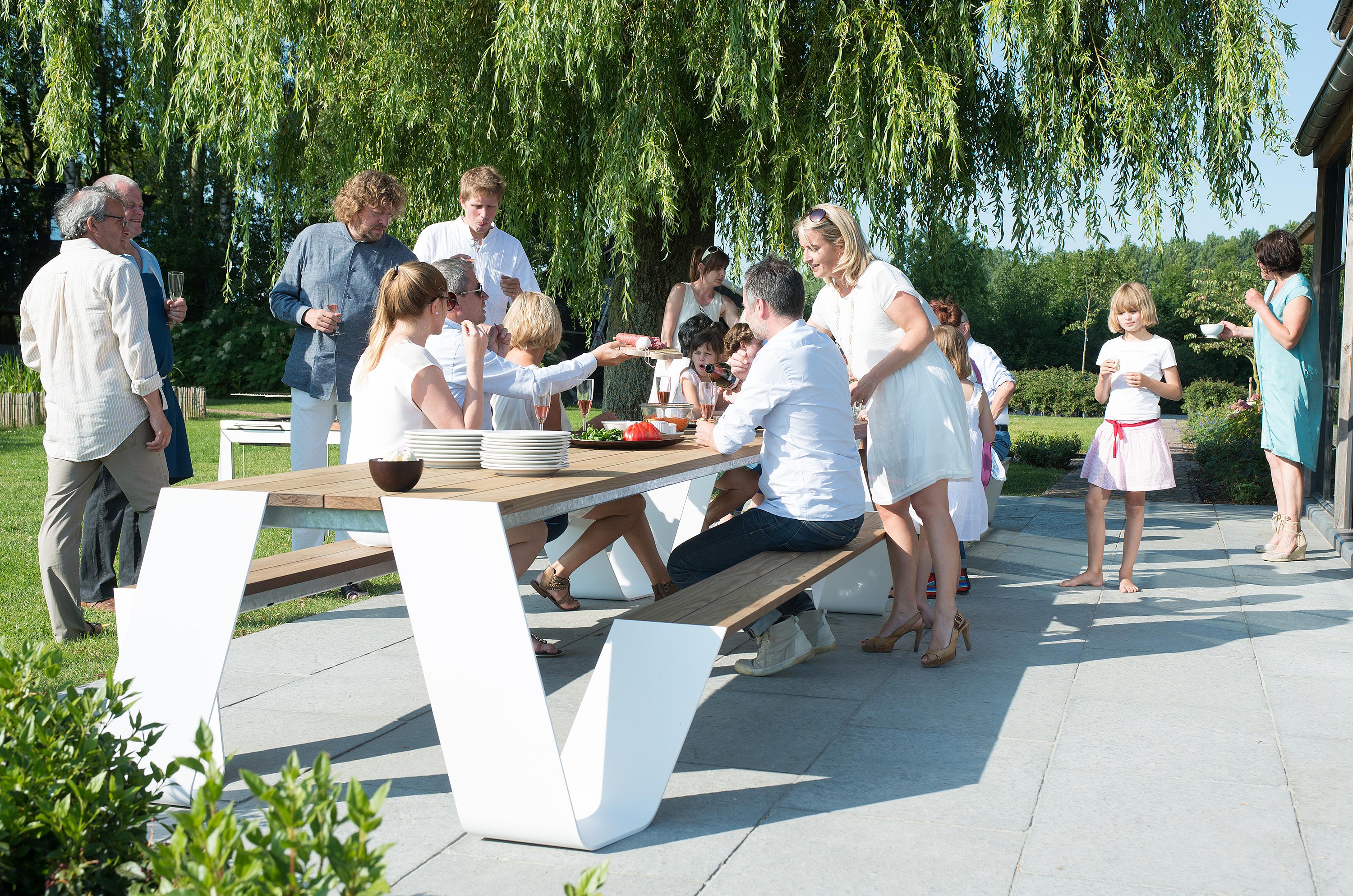 Extended family enjoying a summer barbecue around two Hopper tables on a terrace beside a garden, with adults serving food and children moving around the group.