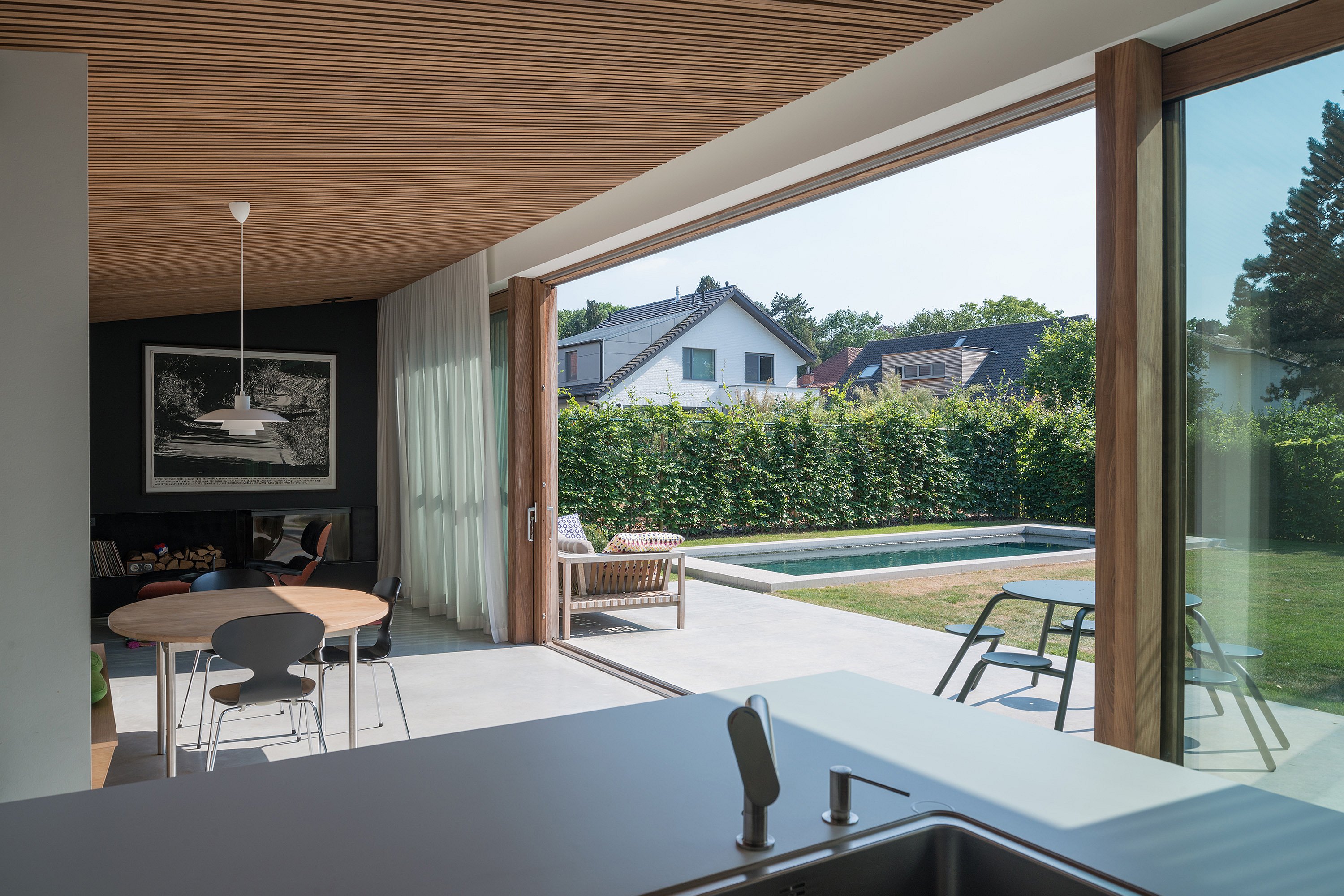 Blick von der Küche durch vollständig geöffnete Glasschiebetüren auf einen minimalistischen Garten mit schmalem Swimmingpool; Im Innenbereich sitzen ein runder Esstisch und eine Pendelleuchte unter einer Holzlattendecke, während der grüne Virus-Tisch draußen auf der Terrasse sichtbar ist.