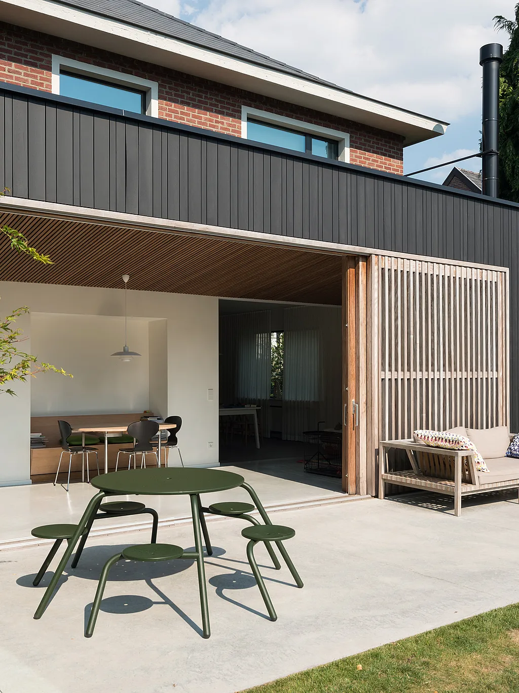 Eigentijds bakstenen huis met een donker geklede tuinuitbreiding die uitkomt op een betonnen terras, waar een groene ronde Virus picknicktafel met vijf bevestigde krukken op de voorgrond staat naast een kussende buitenbank en een grote groenblijvende boom.