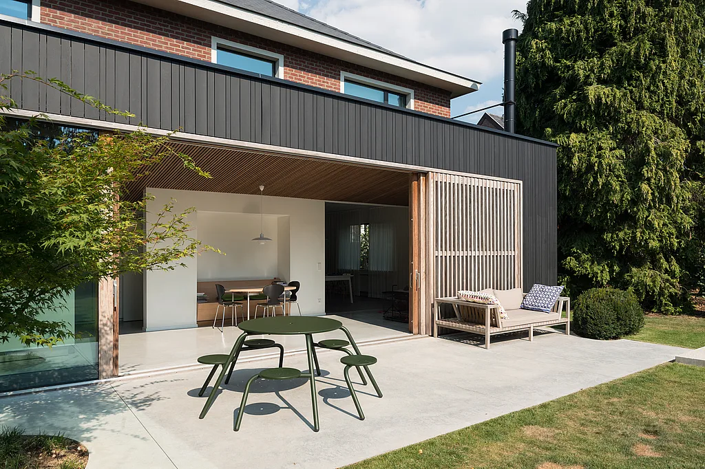 Contemporary brick house with a dark-clad garden extension opening onto a concrete terrace, where a green round Virus picnic table with five attached stools sits in the foreground beside a cushioned outdoor bench and a large evergreen tree.
