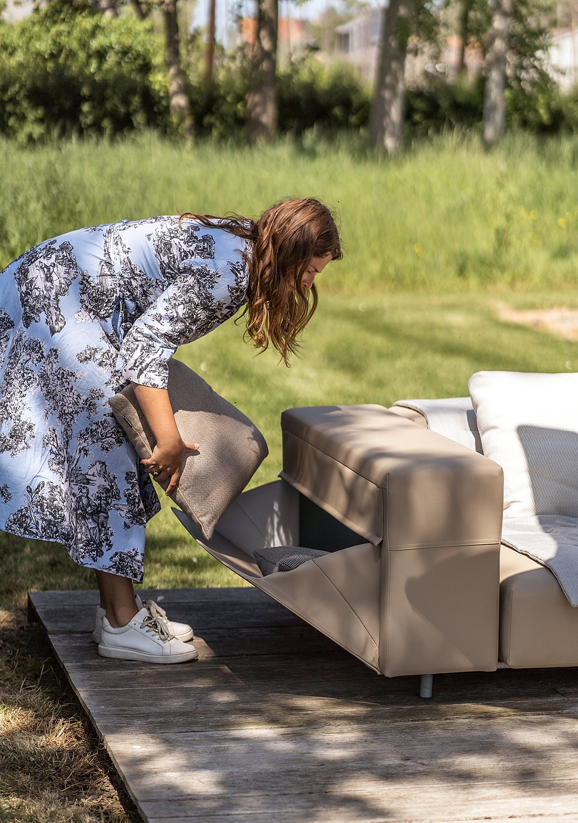 Femme se penchant à côté d’un canapé d’extérieur taupe, ouvrant la poche latérale intégrée et plaçant un coussin à l’intérieur sur une terrasse en bois entourée de verdure.