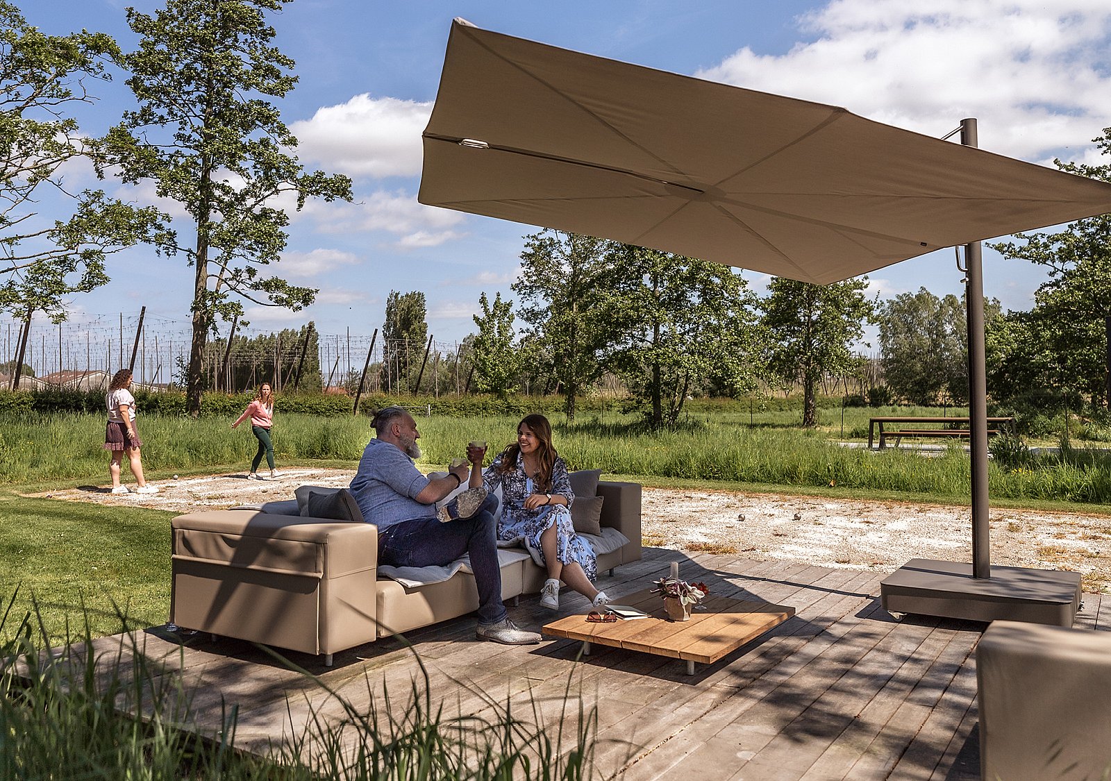 Großer taupefarbener Sensu-Schatten, der über einem niedrigen Außensofa auf einer Holzterrasse auskragt, wo ein Mann und eine Frau neben einem Couchtisch sitzen und sich dahinter ein grasbewachsener Garten und Bäume erstrecken.