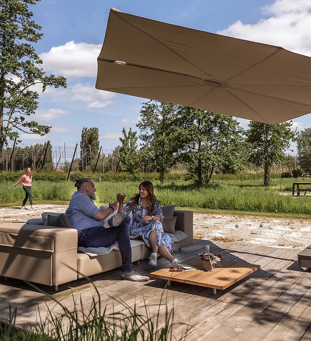 Großer taupefarbener Sensu-Schatten, der über einem niedrigen Außensofa auf einer Holzterrasse auskragt, wo ein Mann und eine Frau neben einem Couchtisch sitzen und sich dahinter ein grasbewachsener Garten und Bäume erstrecken.