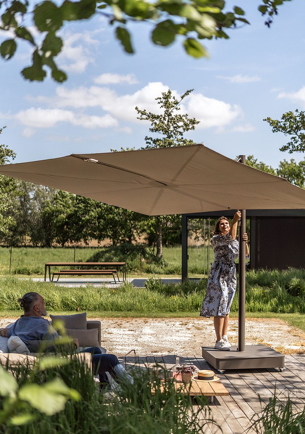 Femme debout sur la base d’un grand store carré Sensu et ajustant le poteau central, tandis qu’un homme se détend sur un canapé extérieur assorti à côté d’une table basse sur une terrasse ensoleillée.