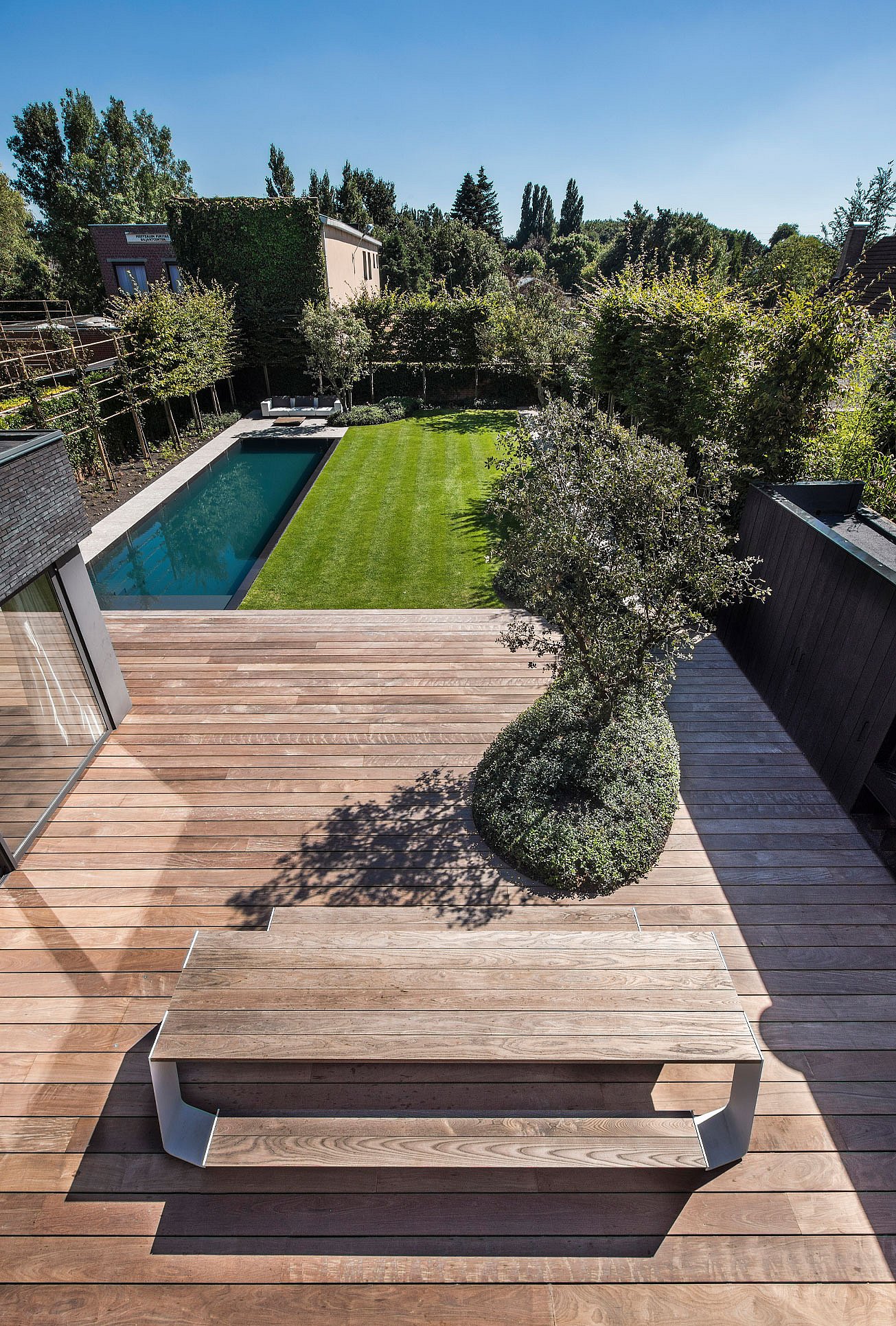 Blick von oben auf eine geräumige Holzterrasse mit einem langen Hopper-Picknicktisch im Vordergrund, die hinunter zu einem Garten mit Pool und gepflegtem Rasen führt.