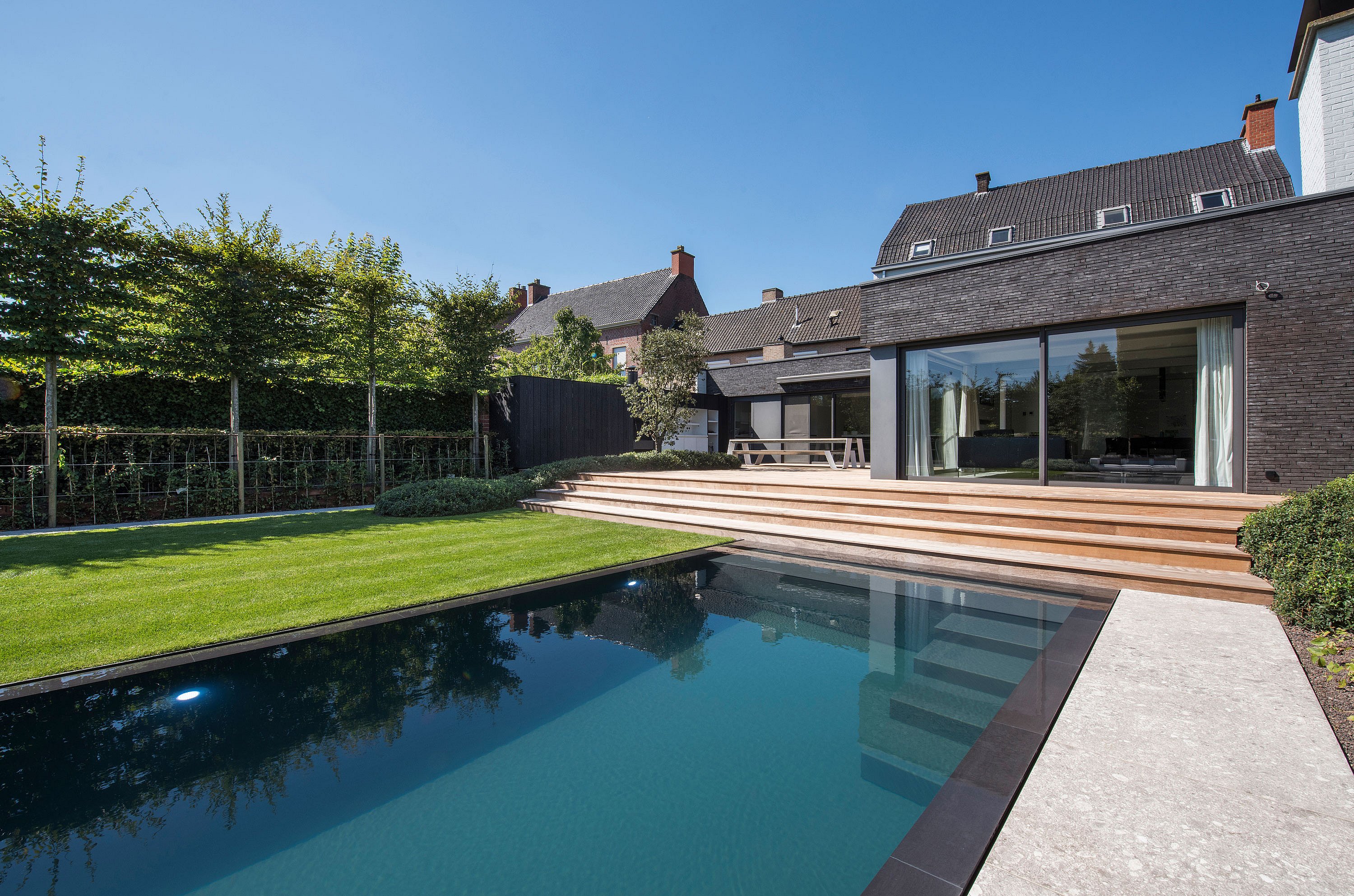   Ein modernes Backsteinhaus mit breiter Holztreppe, die zu einem eleganten Swimmingpool hinabführt. Auf der oberen Terrasse neben großen Glasschiebetüren steht ein Hopper-Picknicktisch.