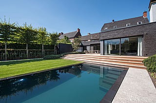 Ein modernes Backsteinhaus mit breiter Holztreppe, die zu einem eleganten Swimmingpool hinabführt. Auf der oberen Terrasse neben großen Glasschiebetüren steht ein Hopper-Picknicktisch.