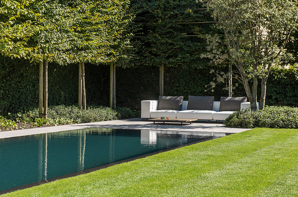 Ein rechteckiger Pool spiegelt die umliegenden Bäume wider. Dahinter, umgeben von üppigem Grün und einem kurzgeschnittenen Rasen, steht auf der Terrasse ein weißes Walrus-Sofa mit niedrigem Couchtisch.