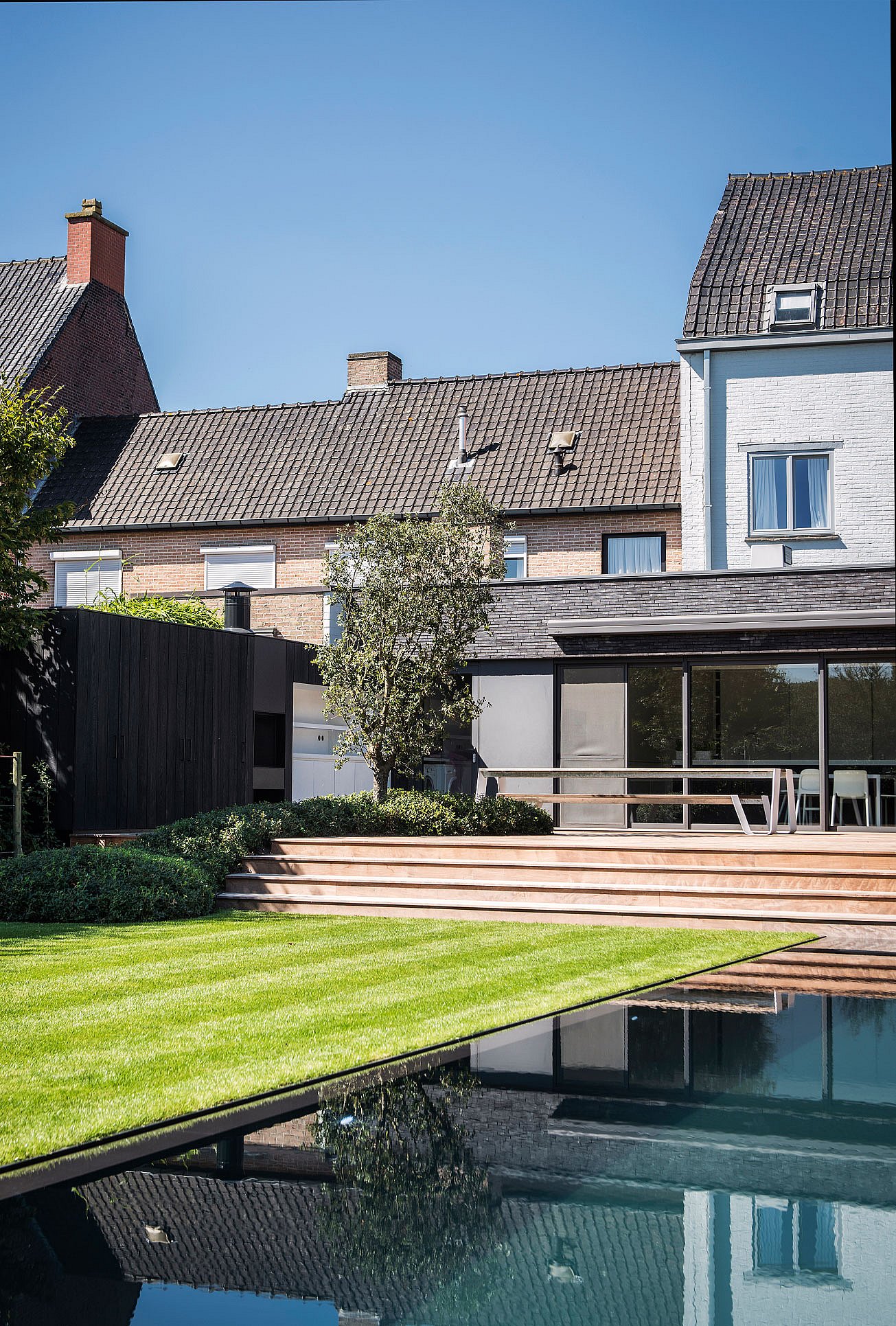 Langer Hopper-Picknicktisch auf einer erhöhten Holzterrasse neben einem modernen Anbau mit Blick auf Pool und Garten, im Hintergrund die benachbarten Backsteinhäuser.
