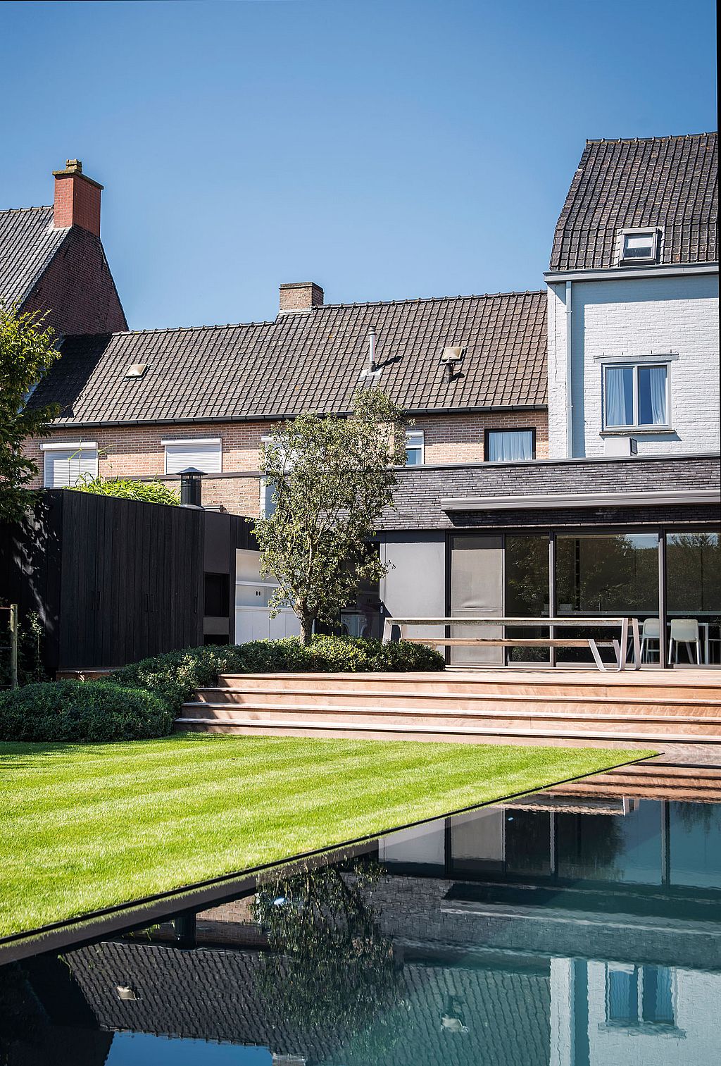 Langer Hopper-Picknicktisch auf einer erhöhten Holzterrasse neben einem modernen Anbau mit Blick auf Pool und Garten, im Hintergrund die benachbarten Backsteinhäuser.
