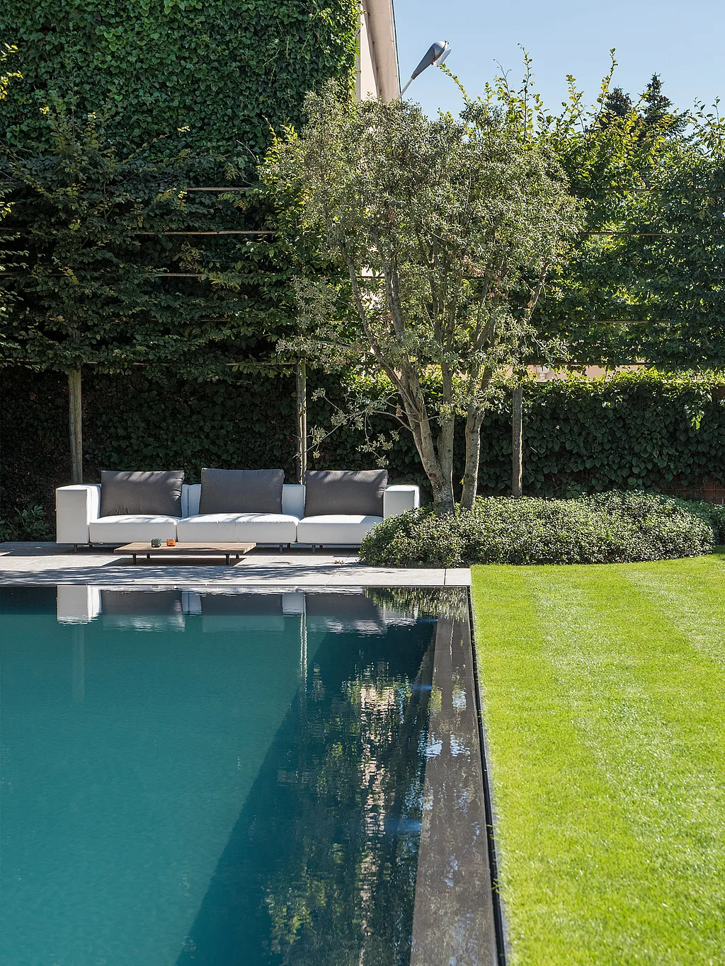 Longue piscine rectangulaire bordée d'une pelouse impeccablement tondue, avec un canapé d'extérieur blanc Walrus et une table basse en bois dissimulés sous des arbres matures au fond du jardin.