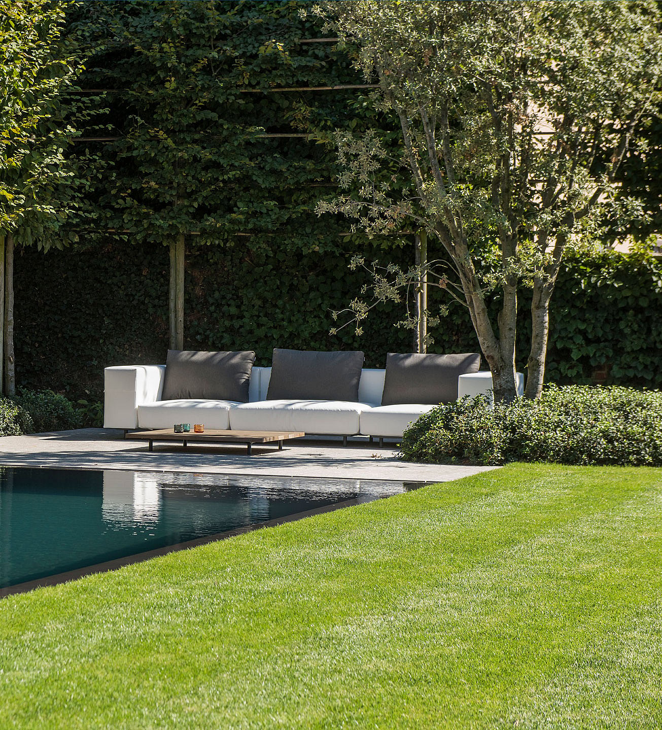   Ein weißes Gartensofa und ein schmaler Holztisch am Pool, eingerahmt von einem leuchtend grünen Rasen im Vordergrund und dichter Bepflanzung im Hintergrund.
