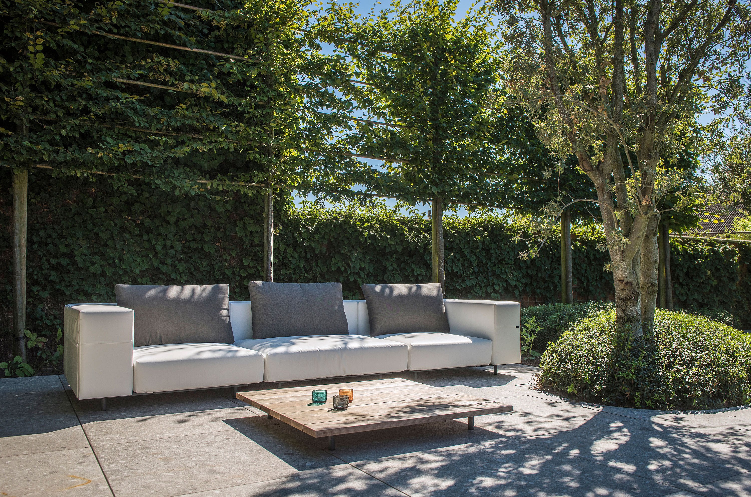   Ein weißes modulares Gartensofa mit dunkelgrauen Rückenkissen und ein niedriger Holztisch auf einer Steinterrasse, umgeben von Bäumen und efeubewachsenen Gartenmauern.