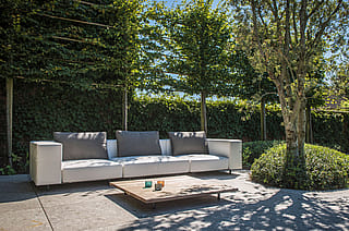 Ein weißes modulares Gartensofa mit dunkelgrauen Rückenkissen und ein niedriger Holztisch auf einer Steinterrasse, umgeben von Bäumen und efeubewachsenen Gartenmauern.