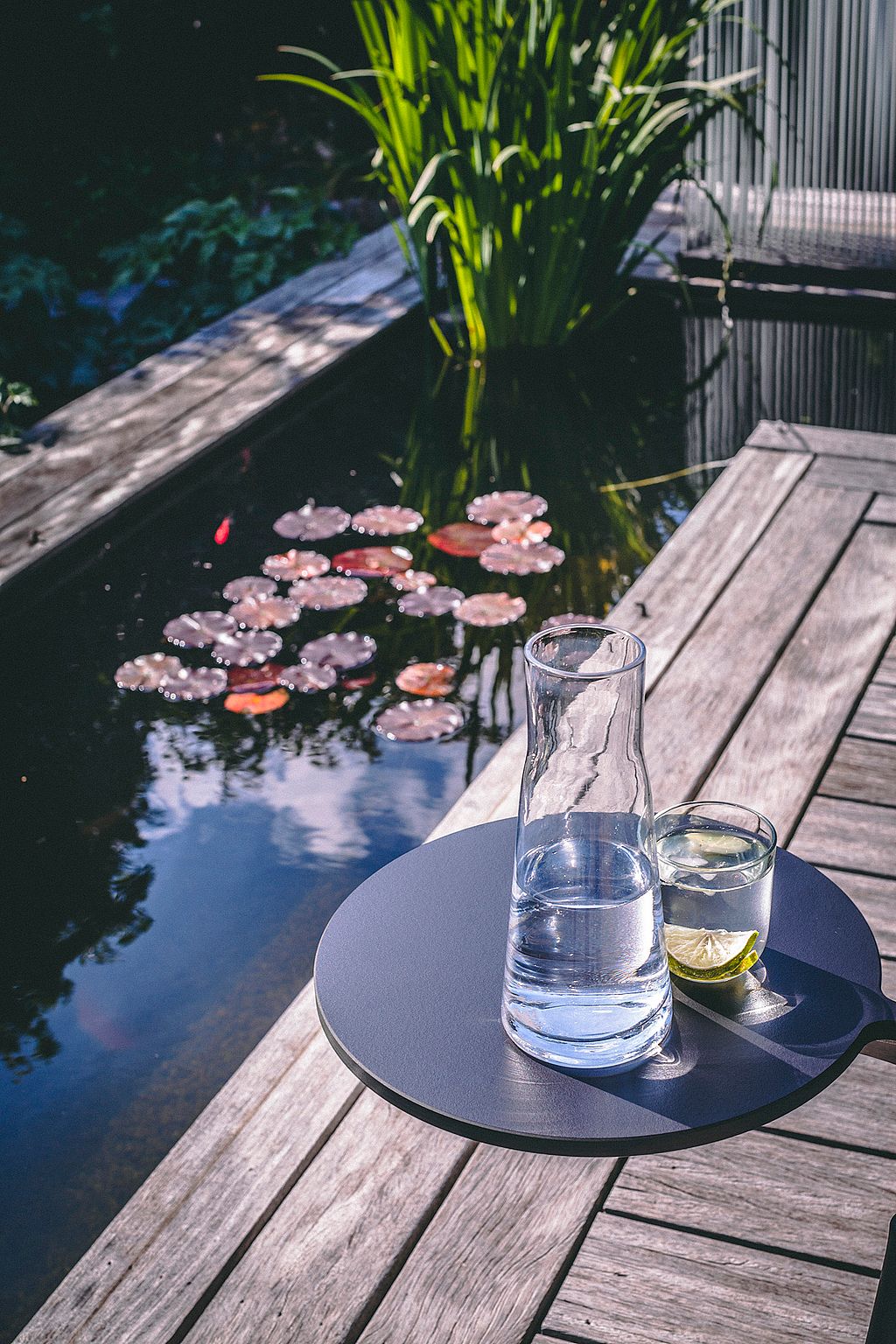 Round dark side table on a wooden terrace holding a glass carafe and a drink with lime, next to a fishpond with lily pads and tall green plants.