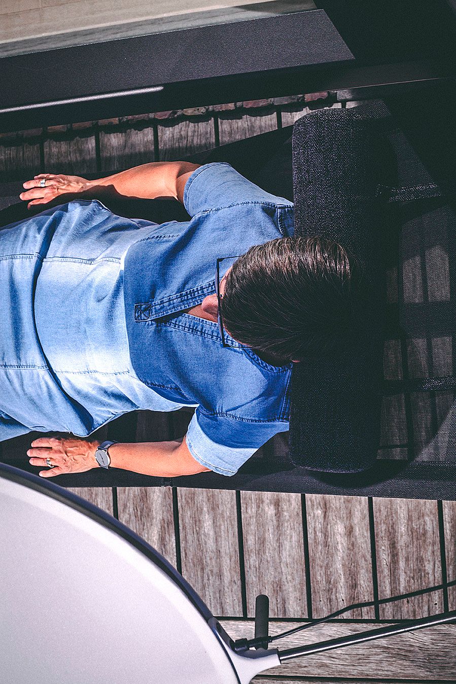 Top view of a person lying back on a dark Sol+Luna outdoor sofa configured as a sunbed on a wooden deck, with the edge of the round shade visible nearby.