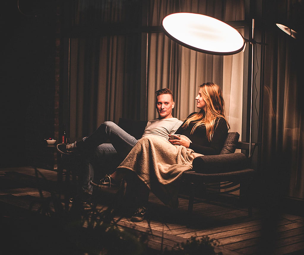 Man and woman relaxing together on a dark outdoor sofa under a glowing round Sol+Luna shade at night, with a blanket over their laps on a cozy wooden patio