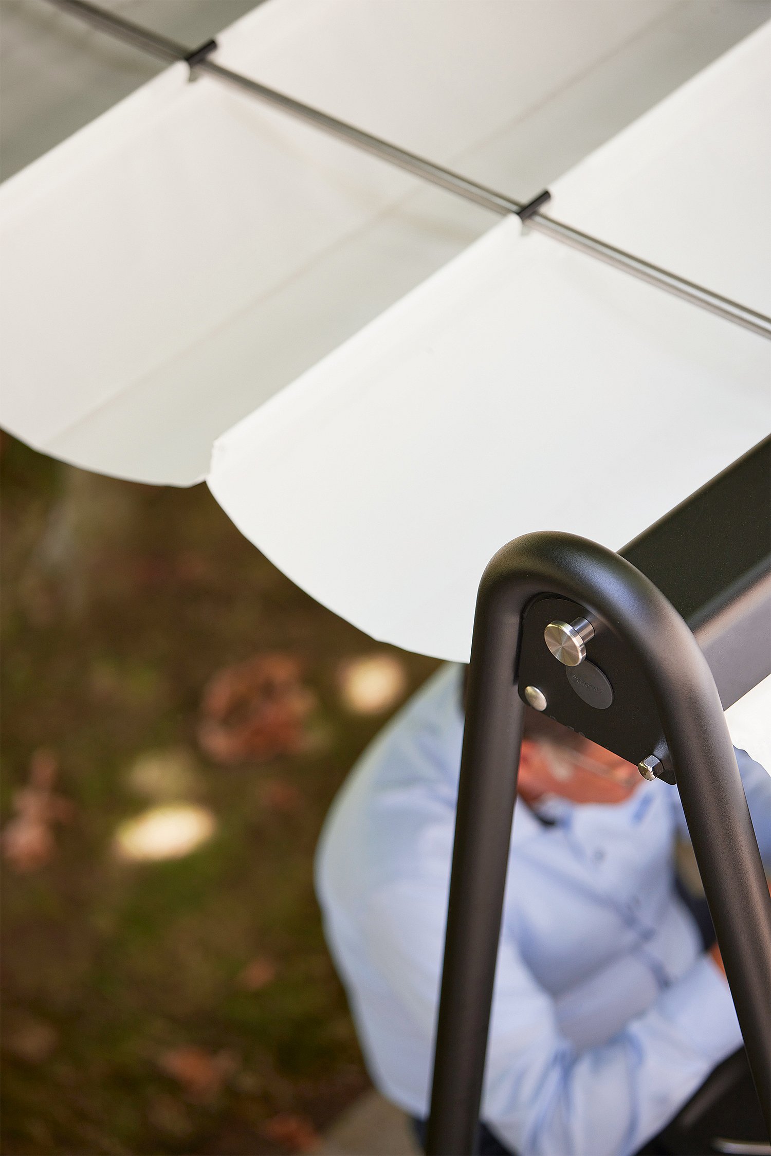 Close-up van het zwarte AMAi-frame en de witte stoffen luifel, met de nadruk op de gebogen ondersteuning en bevestigingsdetails van het buitenkapsysteem.