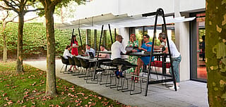 Een groep collega's verzamelde zich rond een zwarte AMAi-tafel onder een witte luifel buiten het kantoor, met laptops, notebooks en drankjes op de tafel onder de bomen.