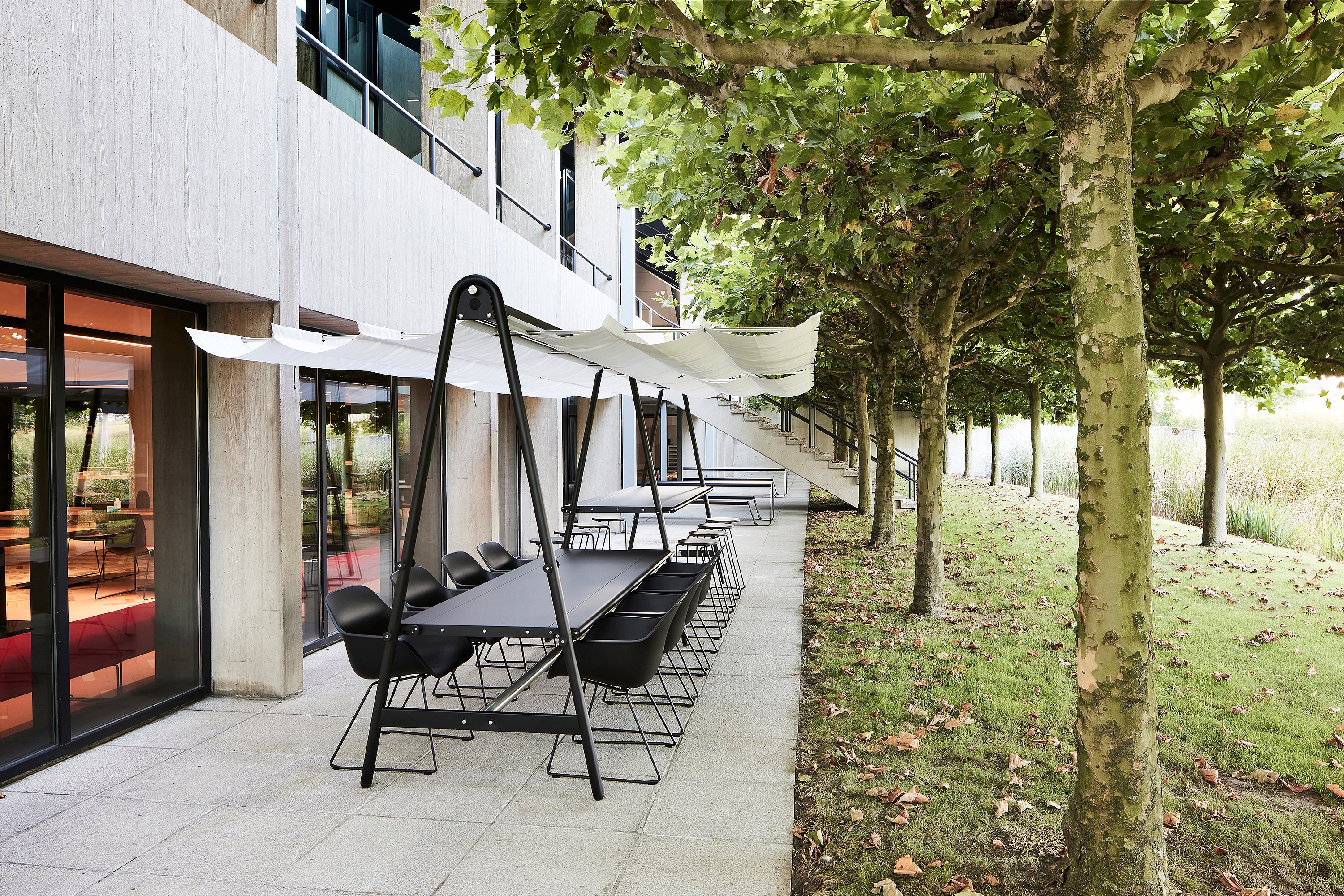 Lange zwarte AMAi vergadertafel voor buiten met een witte stoffen kap en zwarte stoelen naast een modern kantoorgebouw, omzoomd met lommerrijke bomen langs een verhard terras.