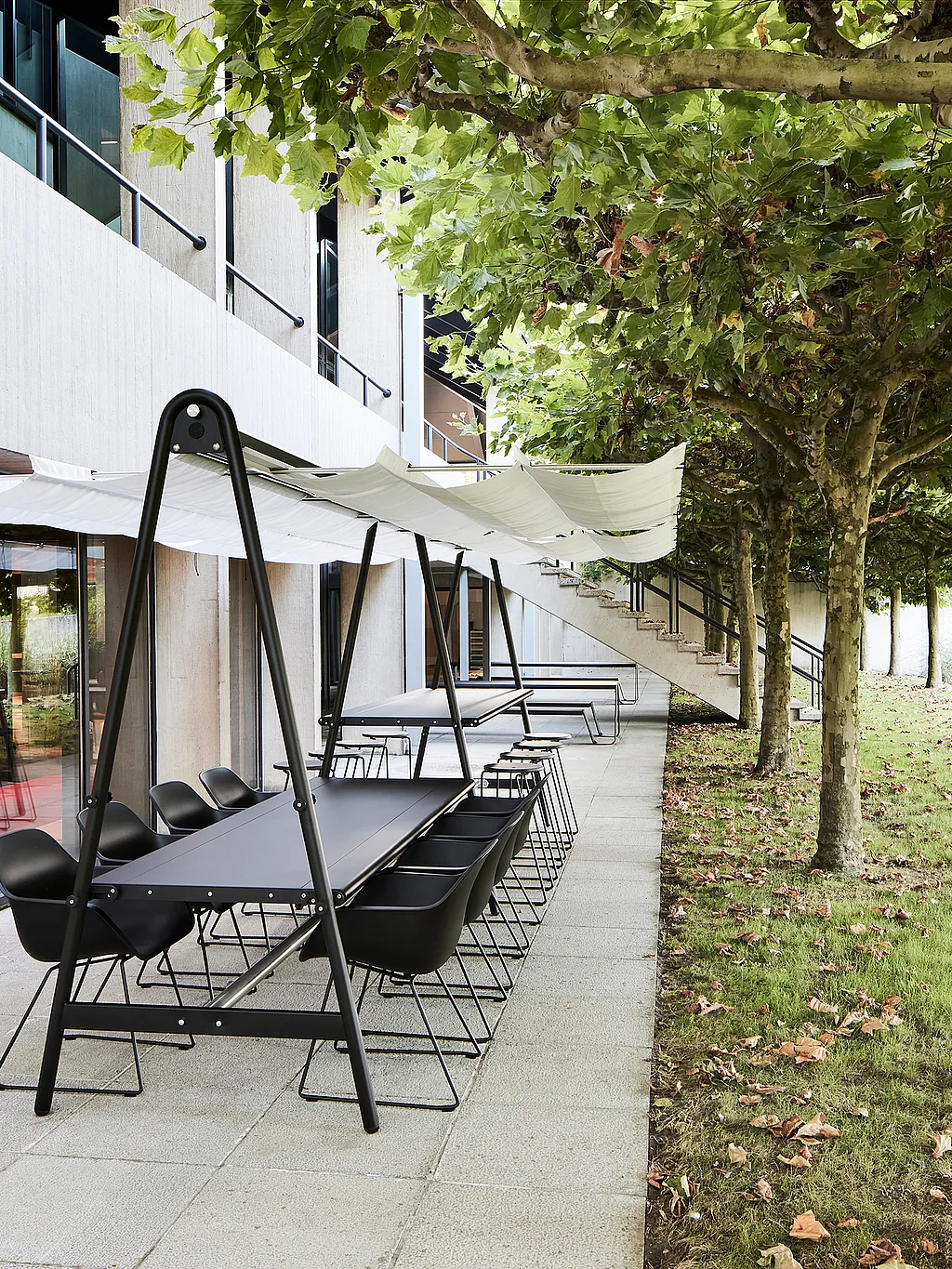 Lange zwarte AMAi vergadertafel voor buiten met een witte stoffen kap en zwarte stoelen naast een modern kantoorgebouw, omzoomd met lommerrijke bomen langs een verhard terras.