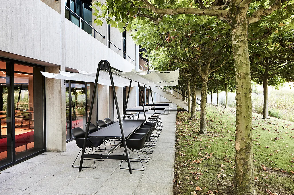 Langer schwarzer AMAi-Tagungstisch im Freien mit weißem Stoffschirm und schwarzen Stühlen neben einem modernen Bürogebäude, gesäumt von grünen Bäumen entlang einer gepflasterten Terrasse.
