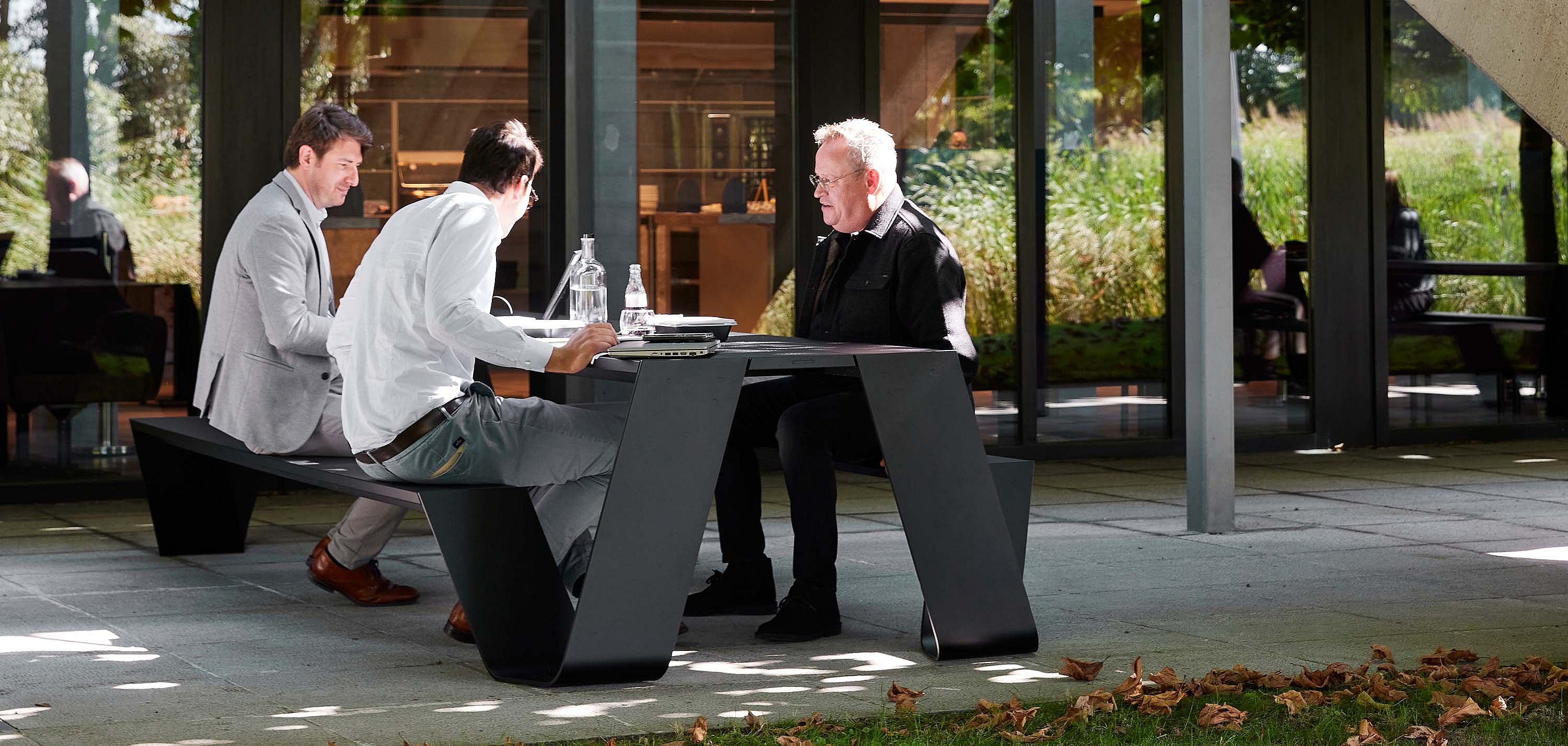 Drie collega's zaten aan een donkere Hopper AA-picknicktafel op een schaduwrijk terras buiten het gebouw en gebruikten de tafel voor een informele buitenbijeenkomst met waterflessen en documenten.