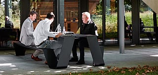 Drie collega's zaten aan een donkere Hopper AA-picknicktafel op een schaduwrijk terras buiten het gebouw en gebruikten de tafel voor een informele buitenbijeenkomst met waterflessen en documenten.